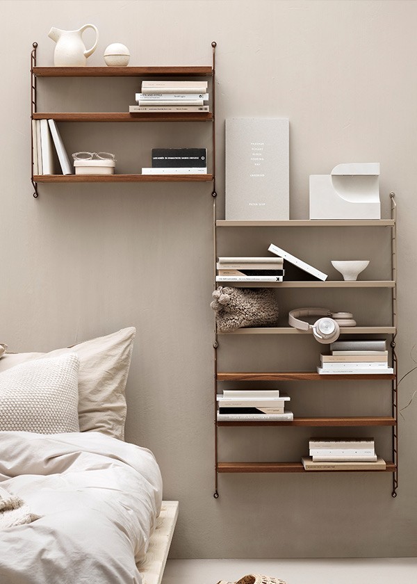 String Pocket shelving in a bedroom, styled with books, ceramics and decorative objects alongside a bed.
