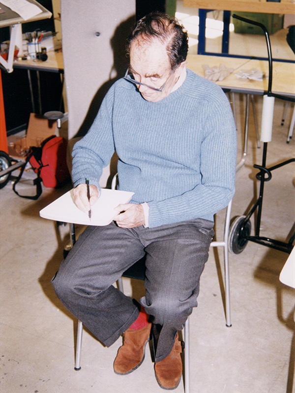 Vico Magistretti sitting on a chair and sketching