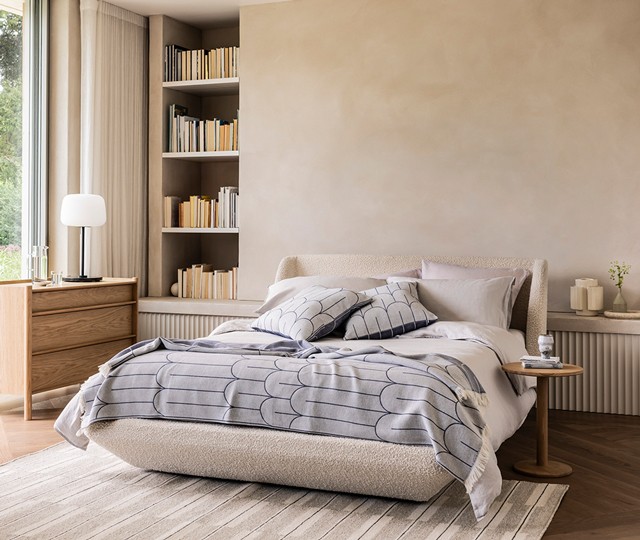 Cream bouclé upholstered platform bed with a grey and blue arched-pattern throw blanket and matching pillows, styled on a striped area rug in a minimalist bedroom.