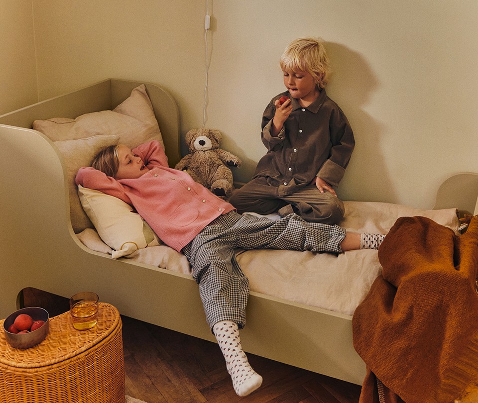 Minimalist olive green wooden toddler bed styled with beige linen bedding, shown with two young children playing and relaxing on the mattress.