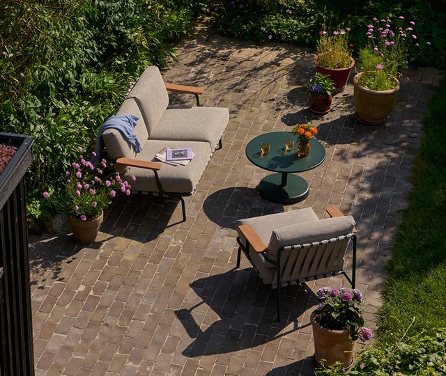 Modern grey outdoor sofa and matching armchair with wood armrests, arranged with a round dark green metal coffee table on a brick patio surrounded by potted flowers.