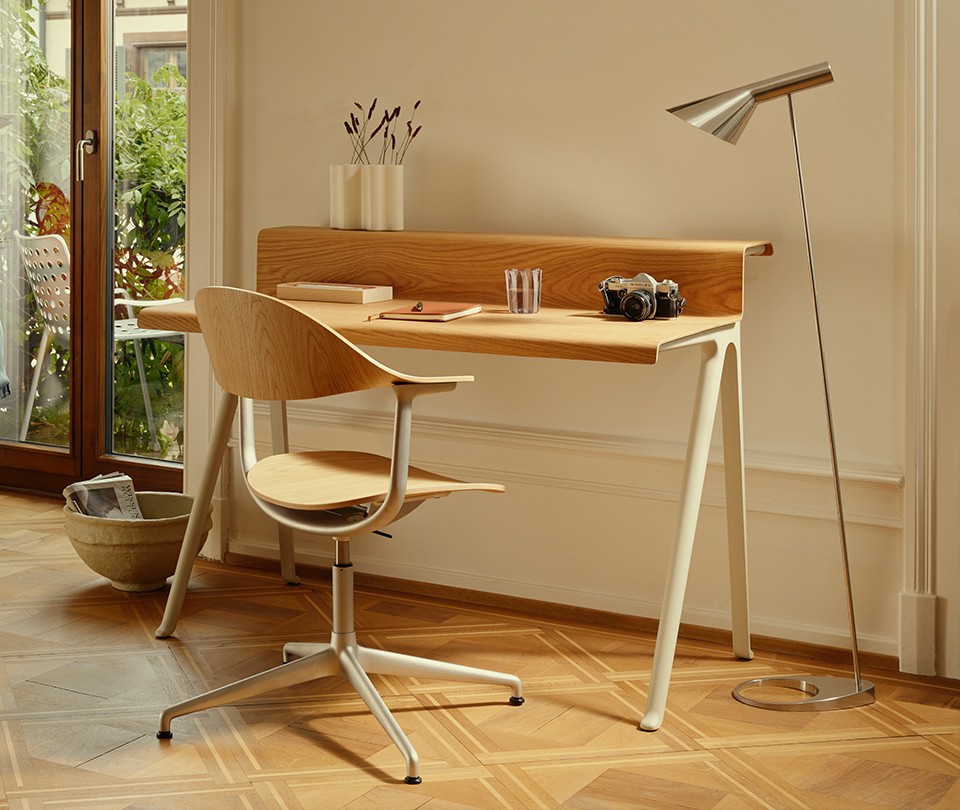 Minimalist oak wood writing desk with cream legs paired with a matching swivel office chair, styled on a parquet wood floor next to a metallic floor lamp.