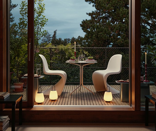 A cozy outdoor balcony dining area with two modern white chairs and a round table set for two, surrounded by plants. Soft lighting comes from two glowing lamps placed on the wooden floor.