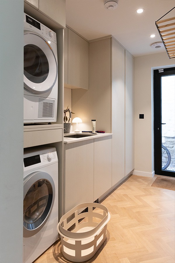 Modern utility room featuring stacked white washer and dryer units, beige fitted cabinets, and herringbone flooring.