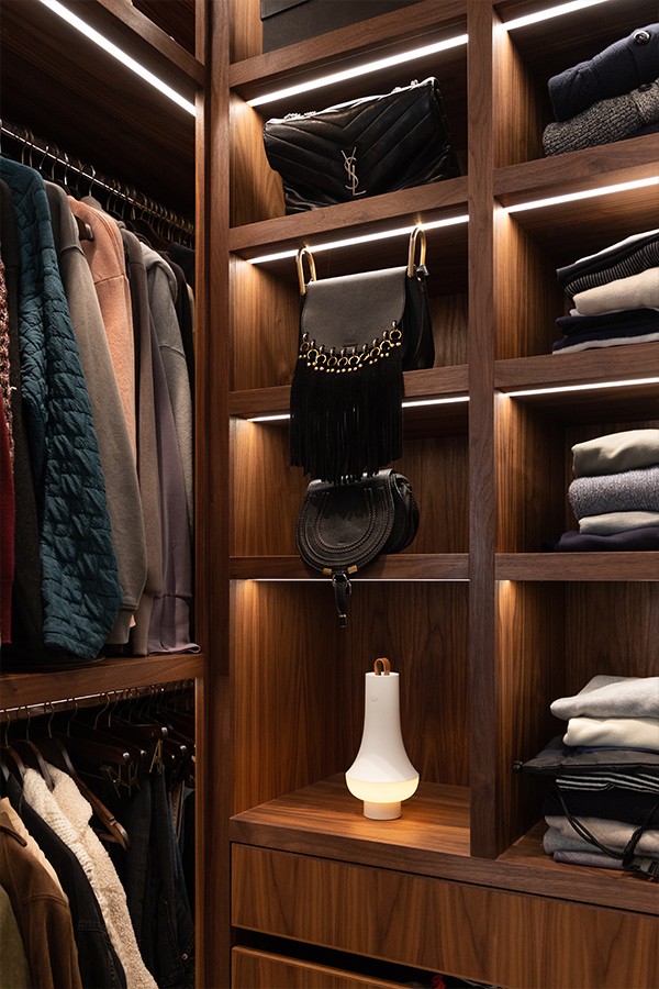 Dark wood walk-in closet with LED-lit shelves, hanging clothes, designer handbags, and a white portable lamp.