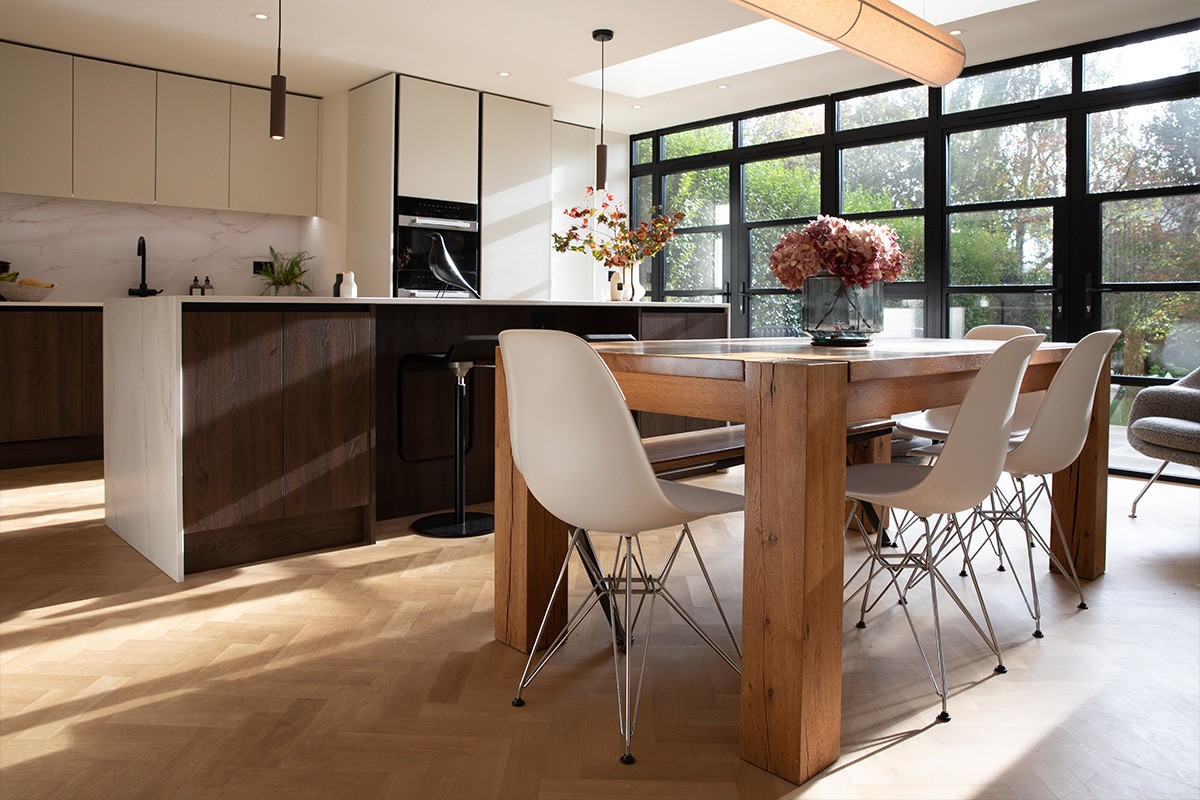 Modern open-plan kitchen and dining room featuring a wooden table, white shell chairs, and a dark wood island.