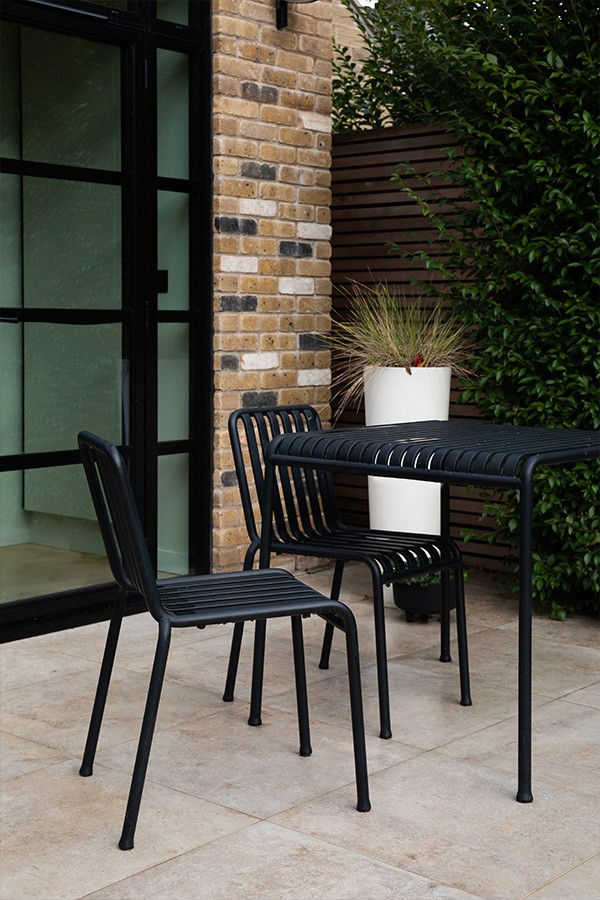 Black slatted metal bistro table and two matching chairs set on a light stone patio next to brick wall.