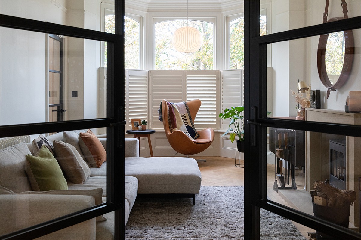 Living room viewed through black-framed glass doors, featuring a grey sectional sofa and a cognac leather egg chair.