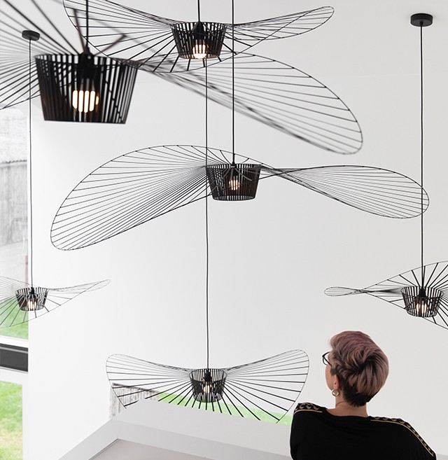 A person with short, light-coloured hair and glasses looks at modern, abstract black wire pendant lights suspended from the ceiling in a bright, minimalist room with large windows.