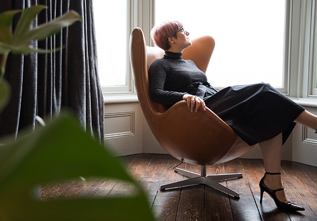 A woman with short hair and glasses sits in a modern brown egg chair by a large window, wearing a black turtleneck, black skirt, and heels. Wooden floors and indoor plants are also visible.