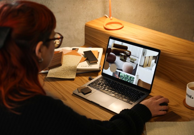 A person with red hair works at a wooden desk, viewing furniture and interior design images on a laptop. Fabric swatches, papers, and a mug of tea are nearby.