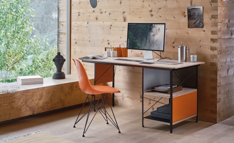 Orange Eames Plastic chair paired with a wooden desk featuring built-in side storage in a wooden room with a large window.
