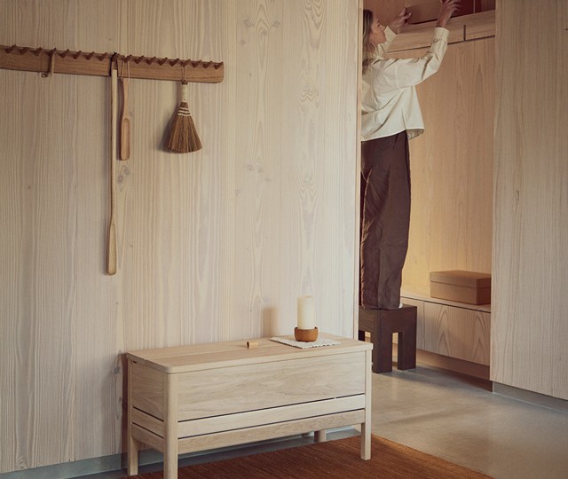 Light oak storage bench set against light wooden paneled walls with a matching wooden peg rack hanging above.
