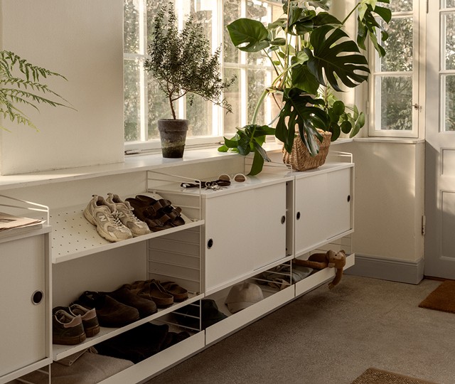 White modular wire shelving system with sliding door cabinets and angled shoe racks, styled with footwear, potted plants, and accessories in a sunlit hallway.