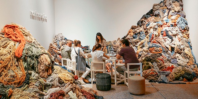 eople sitting at a table surrounded by massive piles of colorful textile fibers and yarn at a Nanimarquina exhibit.