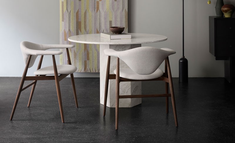 A round marble table with a book and bowl on top, flanked by two modern chairs with wooden legs, set on a dark floor in a minimalist room