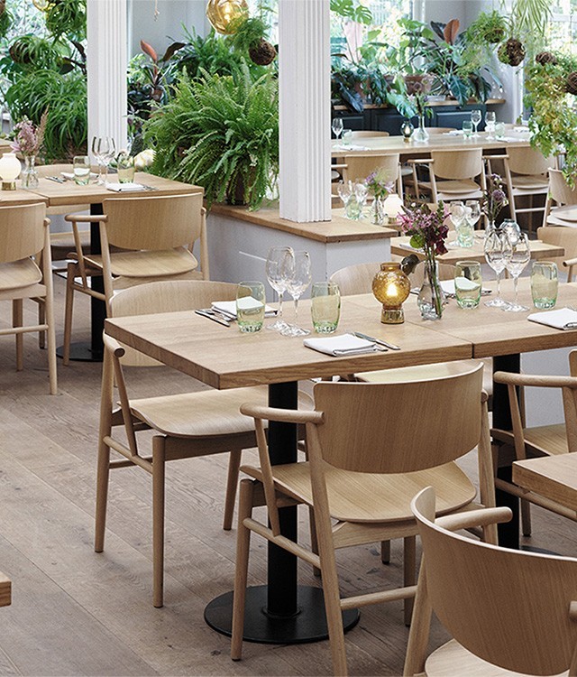 Modern restaurant with wooden tables and chairs, neatly set with glasses and napkins. Large windows and abundant green plants create a bright, airy, natural atmosphere. Decorative lights hang from the ceiling.