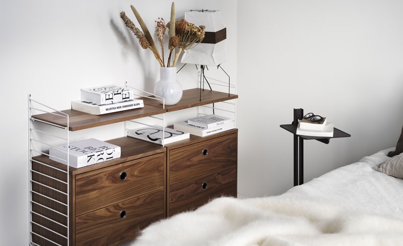 Wall-mounted walnut drawer unit with white wire side panels set beside a bed with fluffy white blanket.